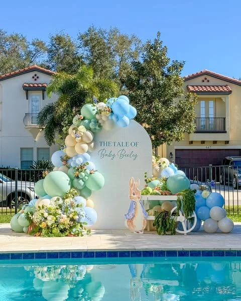 Peter Rabbit themed baby shower backdrop with a pastel balloon garland and character cutout by a pool.