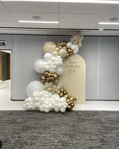 Modern retirement party decor featuring a personalized beige backdrop and a white and gold balloon garland.
