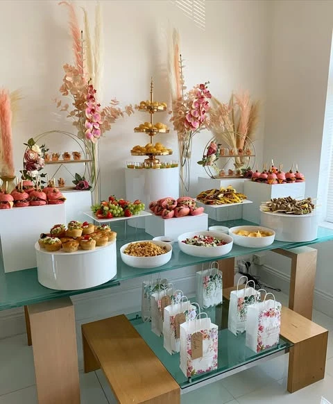 A baby shower food table setup using white risers and a gold tiered stand to display a variety of bite-sized foods.