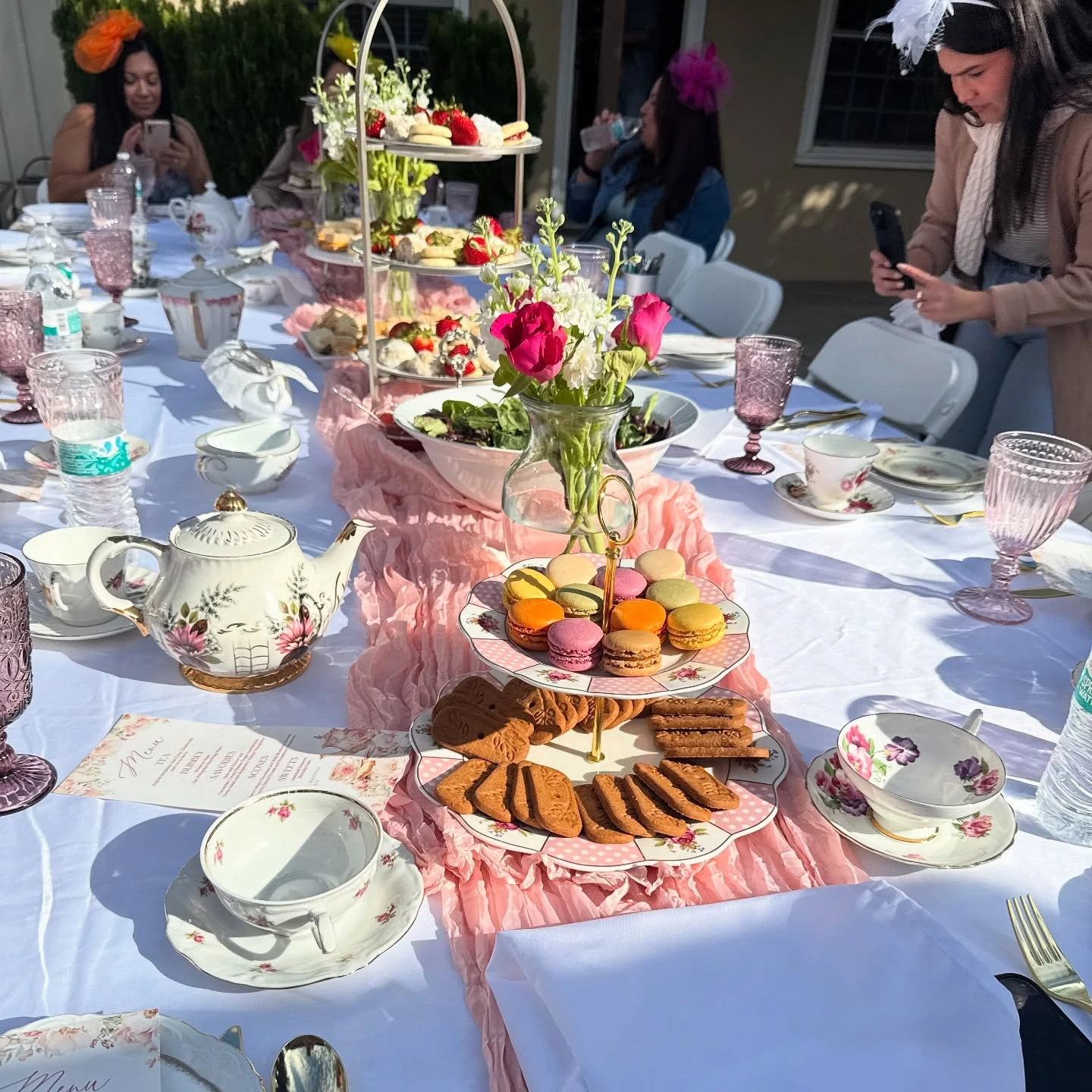 A long white table set for a tea party bridal shower with floral teacups, a pink runner, and tiered stands holding macarons and desserts.
