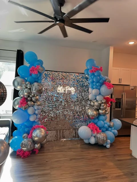 A Mamma Mia themed party photo backdrop with a silver sequin wall and blue and pink balloon garlands.