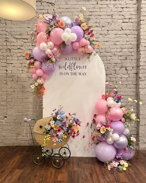 A wildflower baby shower photo backdrop featuring a white arched sign, a pastel balloon garland with flowers, and a vintage wicker pram filled with a floral arrangement.