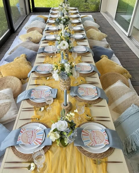 Long picnic table set for a 'Here Comes the Sun' baby shower with yellow and blue decor and floor cushions.