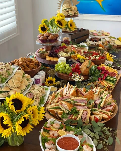 A long wooden table covered in a large grazing spread for a 'You Are My Sunshine' baby shower, with large sunflower bouquets.