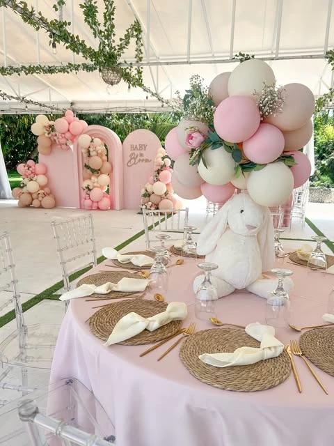 A white plush bunny baby shower centerpiece on a pink tablecloth, with a cloud of pink and white balloons attached above it.
