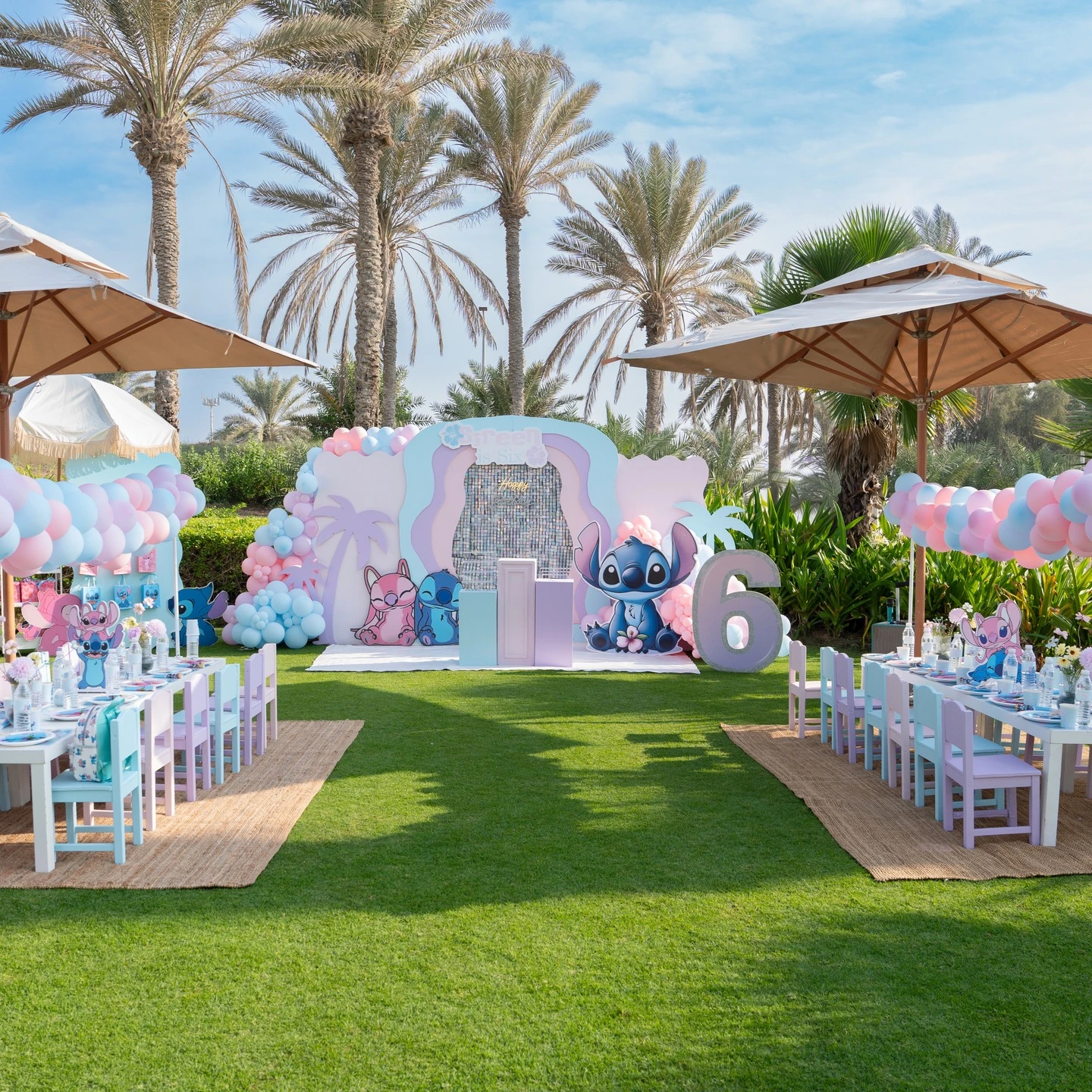 Wide view of an outdoor Stitch and Angel birthday party with pastel decorations, a large photo backdrop, and two long kids' tables.