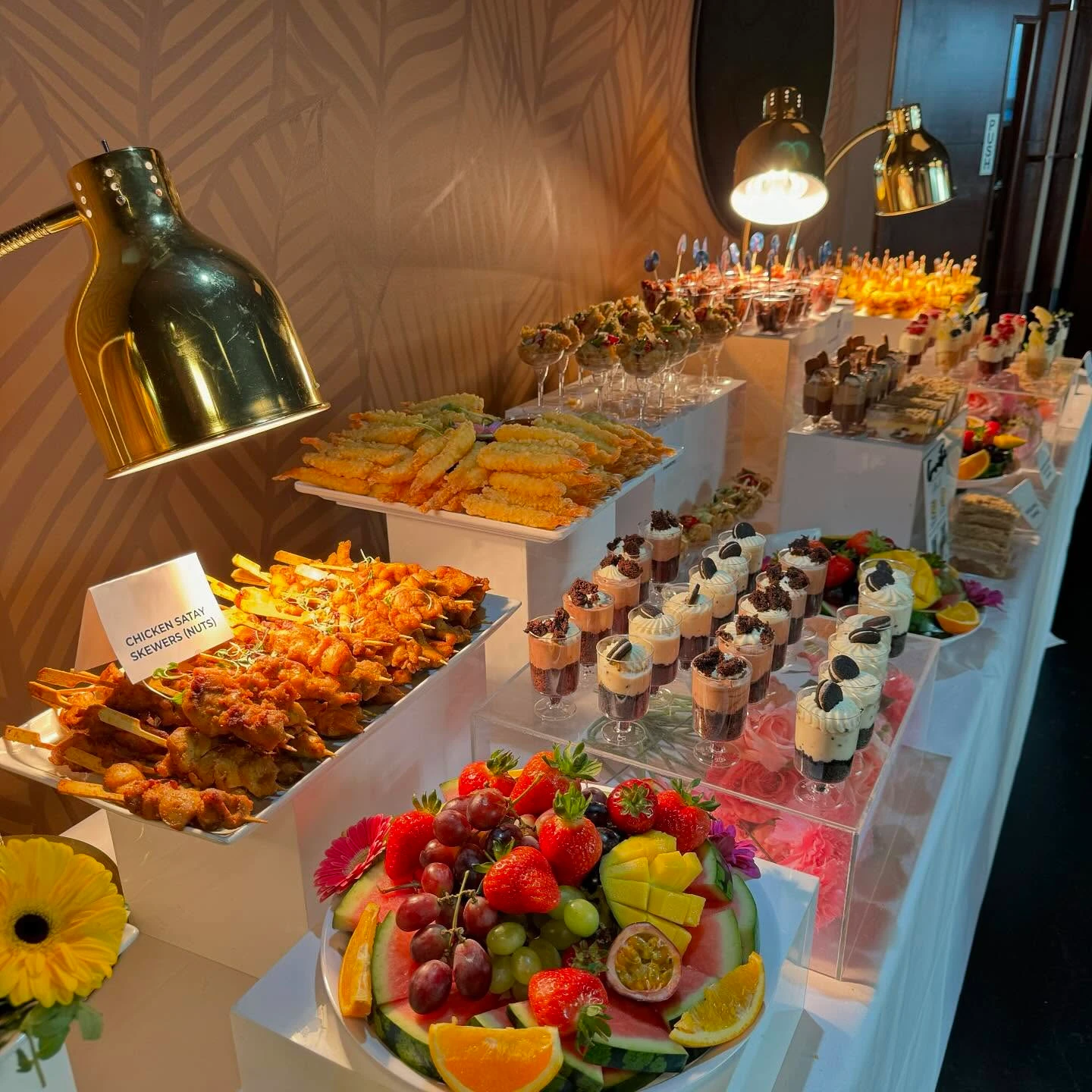 Elaborate baby shower food table setup featuring tiered displays, a heat lamp over chicken skewers, and individual dessert cups.