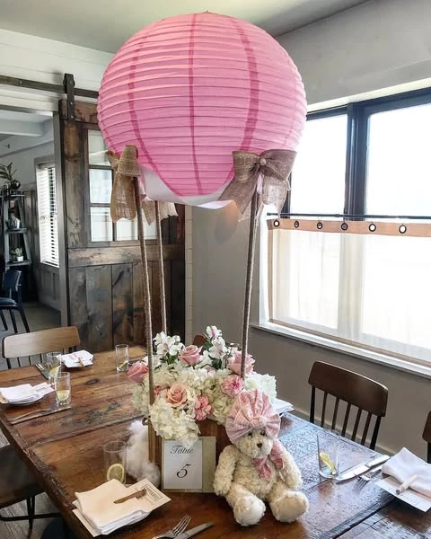 A DIY hot air balloon baby shower centerpiece on a wooden table, featuring a pink paper lantern as the balloon and a flower-filled wooden box as the basket, with a small teddy bear