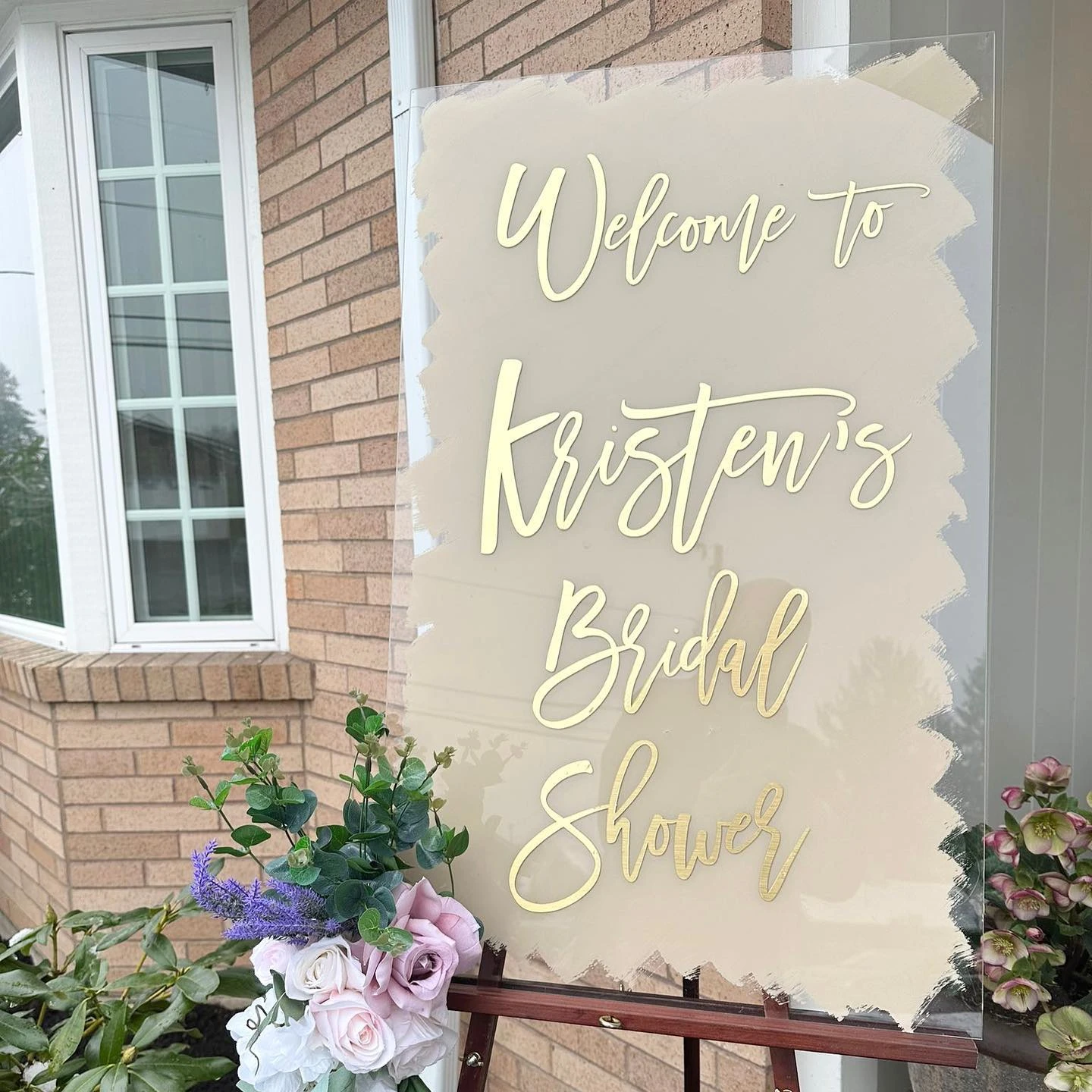 An elegant acrylic welcome sign for a bridal shower, with gold lettering and a floral arrangement.