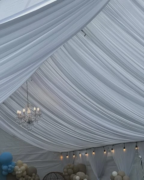 White fabric draping lines the ceiling of an event tent with a chandelier and string lights.