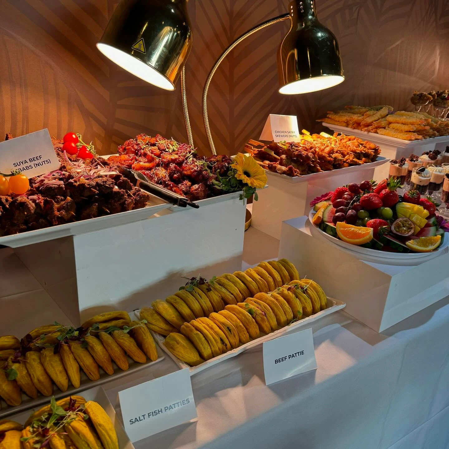 A buffet table with two heat lamps warming platters of beef kebabs and chicken skewers, with food labels for patties in the foreground.