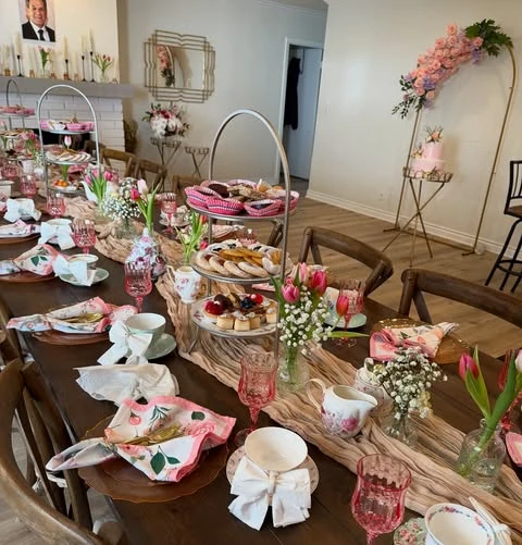 A long wooden table set for a tea party bridal shower with a pink runner, tiered dessert stands, and floral teacups.