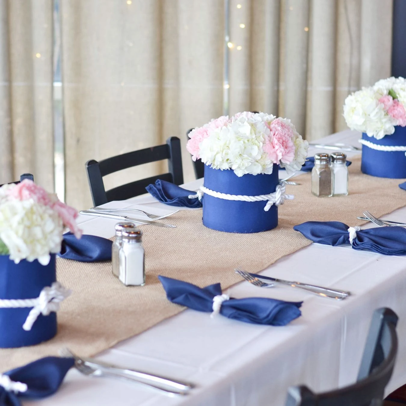 A navy blue and white nautical-themed floral centerpiece with pink and white flowers sits on a burlap table runner.
