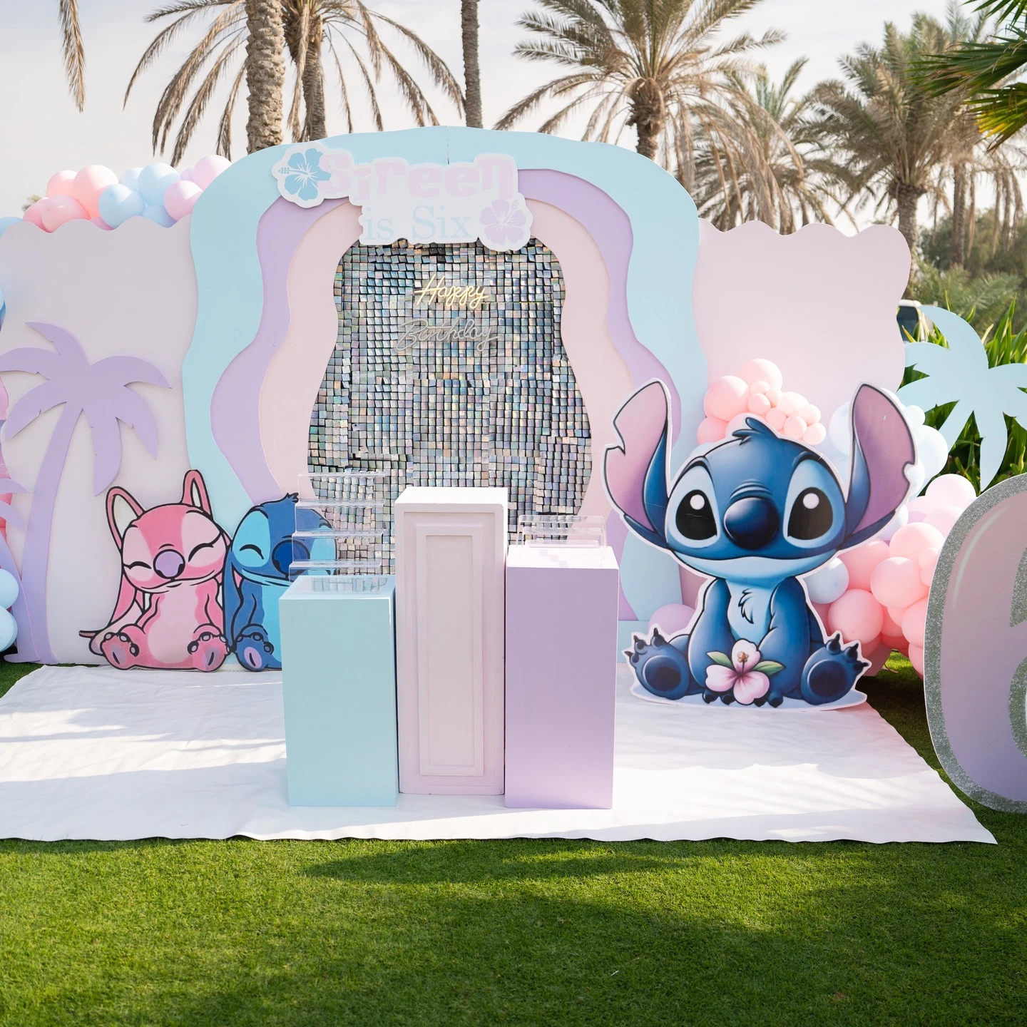 A large pastel-colored party backdrop with cutouts of Stitch and Angel and a silver sequin panel in the middle.