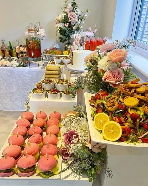 An abundant spring bridal shower buffet with pink mini-burgers, salads, and floral arrangements on tiered white platters.
