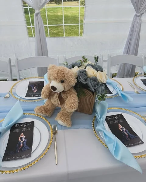 A baby shower table setting with a gold-beaded charger plate, a light blue napkin, and a centerpiece featuring a small teddy bear next to dusty blue and white roses.