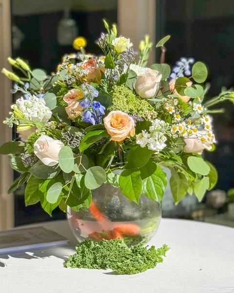 Peter Rabbit themed table centerpiece with fresh carrots submerged in a clear glass bowl vase under a bouquet of roses and wildflowers.