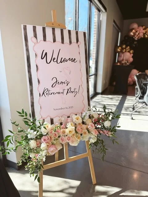 A retirement party welcome sign on an easel, decorated with a large pink and white floral arrangement.