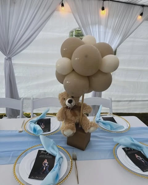 A baby shower centerpiece featuring a teddy bear sitting on a brown box, holding a cluster of tan and cream balloons to look like a hot air balloon.