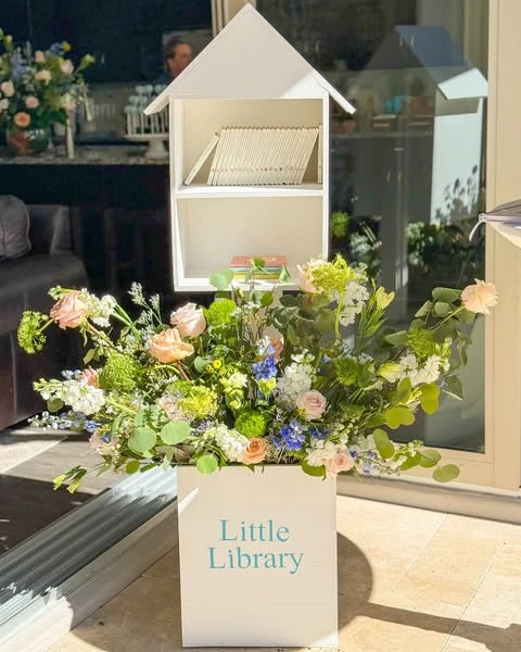 A white house-shaped book drop-off station decorated with a large floral arrangement for a Peter Rabbit baby shower.