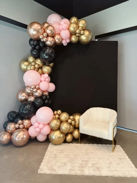 A retirement party photo backdrop with a black wall, a balloon arch in pink, black, and gold, and a white chair.