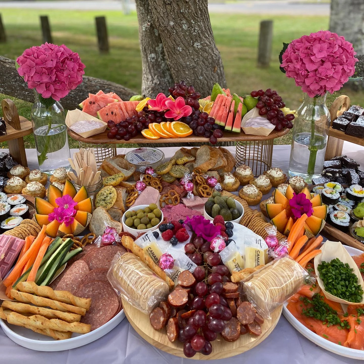 An abundant baby shower grazing table setup featuring platters of fruit, cheese, meats, sushi, and desserts on various stands.