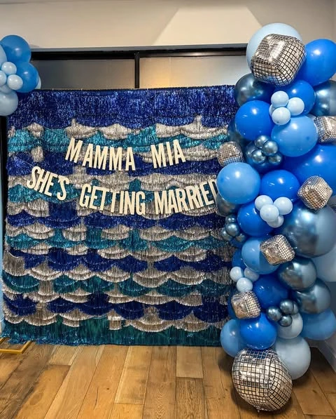 Mamma Mia themed bachelorette party photo backdrop with blue and silver tinsel and a balloon garland with disco balls.