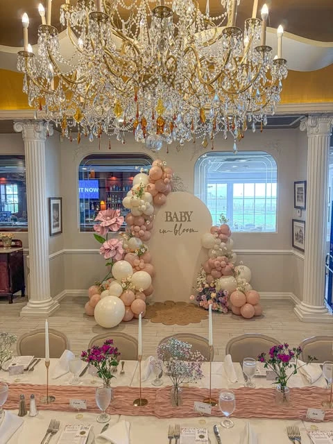 A 'Baby in Bloom' baby shower backdrop with a pink and white balloon arch, seen from across a dining table.