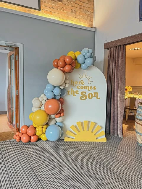 A 'Here comes the Son' baby shower backdrop with a retro balloon garland in terracotta, dusty blue, and mustard yellow.