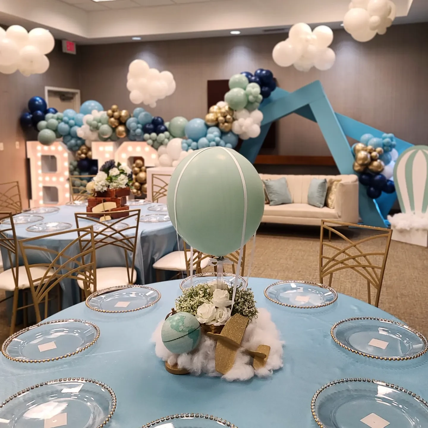 A travel-themed baby shower centerpiece on a blue tablecloth, featuring a mint-green hot air balloon above a basket of flowers resting on a cotton cloud with a miniature globe and