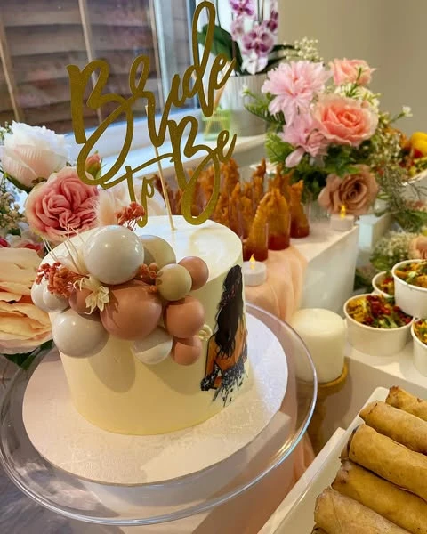 A white bridal shower cake with a gold 'Bride to be' topper and decorative spheres, surrounded by flowers.