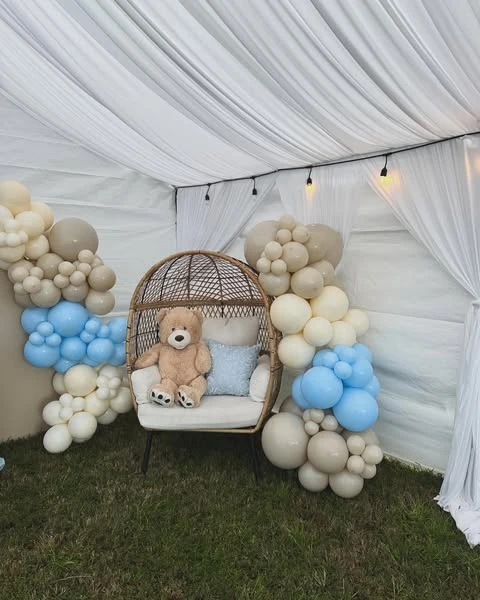 A wicker chair with a teddy bear, framed by blue, tan, and cream balloon garlands, set up as a baby shower photo spot.