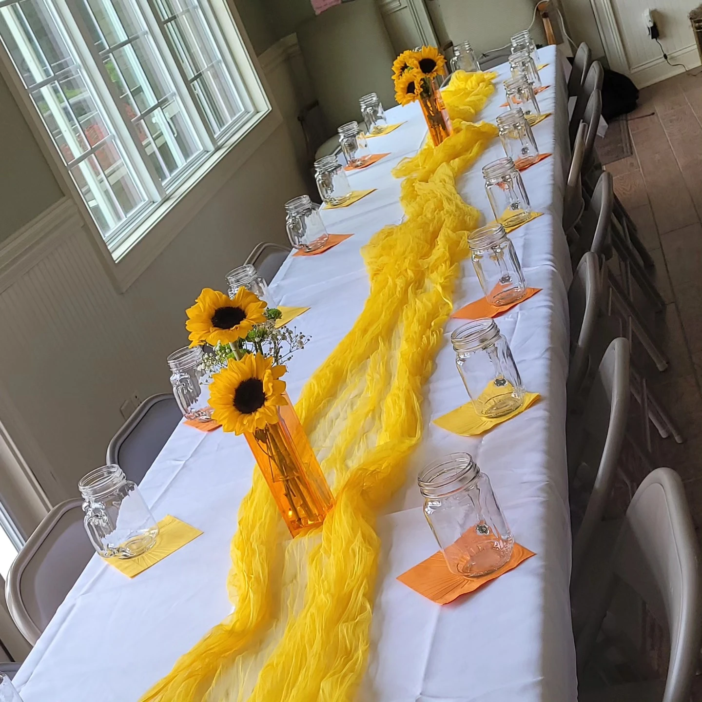 A long baby shower table decorated with a crinkled yellow runner, mason jars, and low sunflower centerpieces in orange vases.