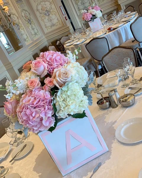 A baby shower centerpiece featuring a white block vase with a pink letter 'A', filled with pink and white roses and hydrangeas on a guest table.