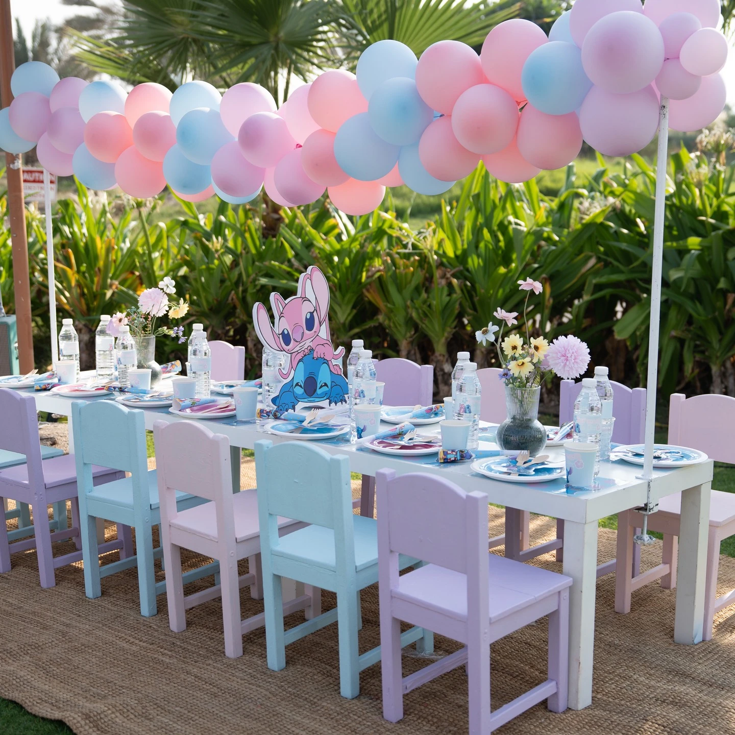 A long kids' party table outdoors with pastel chairs and a Stitch and Angel centerpiece, with a matching pastel balloon garland hanging over it like a canopy.