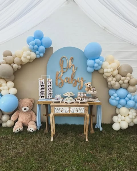 A baby shower dessert station with a blue arched backdrop, a wooden 'Oh Baby' sign, and a balloon garland in blue, tan, and cream. A large teddy bear sits on the grass to the left.