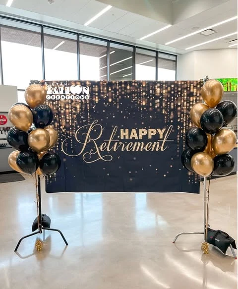 A 'Happy Retirement' photo backdrop in black and gold, framed by two balloon columns on weighted stands.