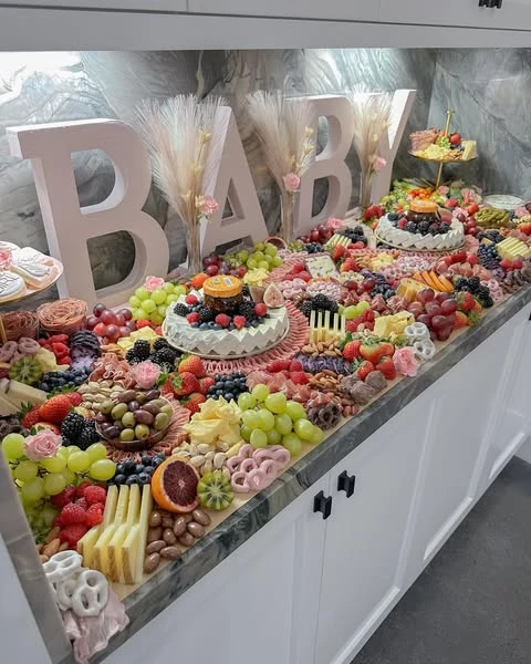 An abundant baby shower grazing table covering a kitchen counter, filled with a dense arrangement of fruits, cheeses, meats, and desserts.