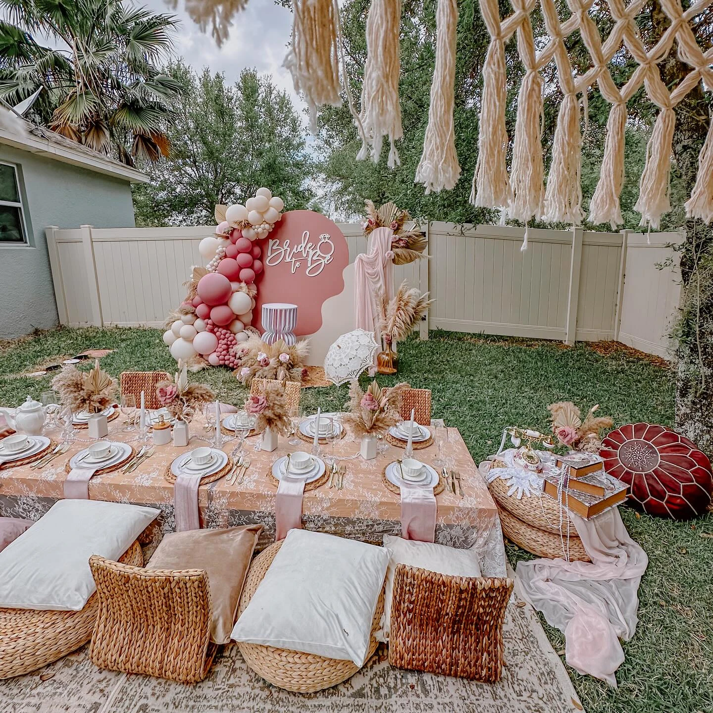 An outdoor tea party picnic setup with macrame hanging overhead and varied floor cushions.
