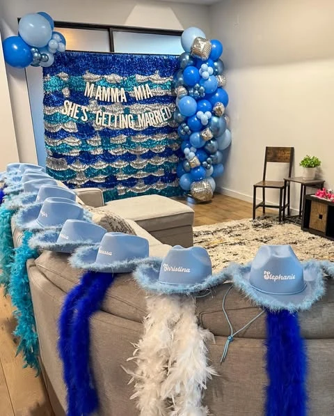 Personalized light blue cowboy hats with feather boas for Mamma Mia bachelorette party guests, lined up on the back of a couch.