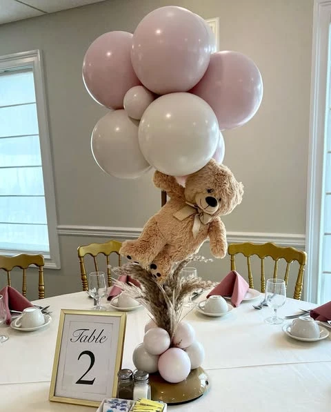 A tall baby shower centerpiece on a table featuring a teddy bear appearing to float beneath a cluster of pink and white balloons.
