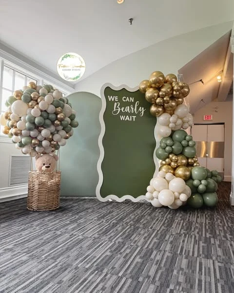 Full view of a 'We Can Bearly Wait' baby shower backdrop featuring a wavy, sage green panel framed by a sage green, white, and chrome gold balloon garland.