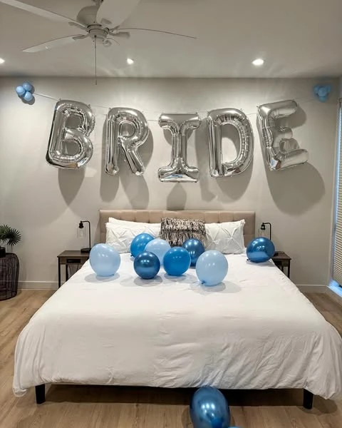 A bedroom decorated for a bachelorette party with large silver 'BRIDE' foil balloons on the wall and blue balloons on the bed.