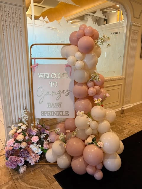 A baby shower welcome sign on a gold stand, decorated with a cascade of pink and white balloons and flowers.