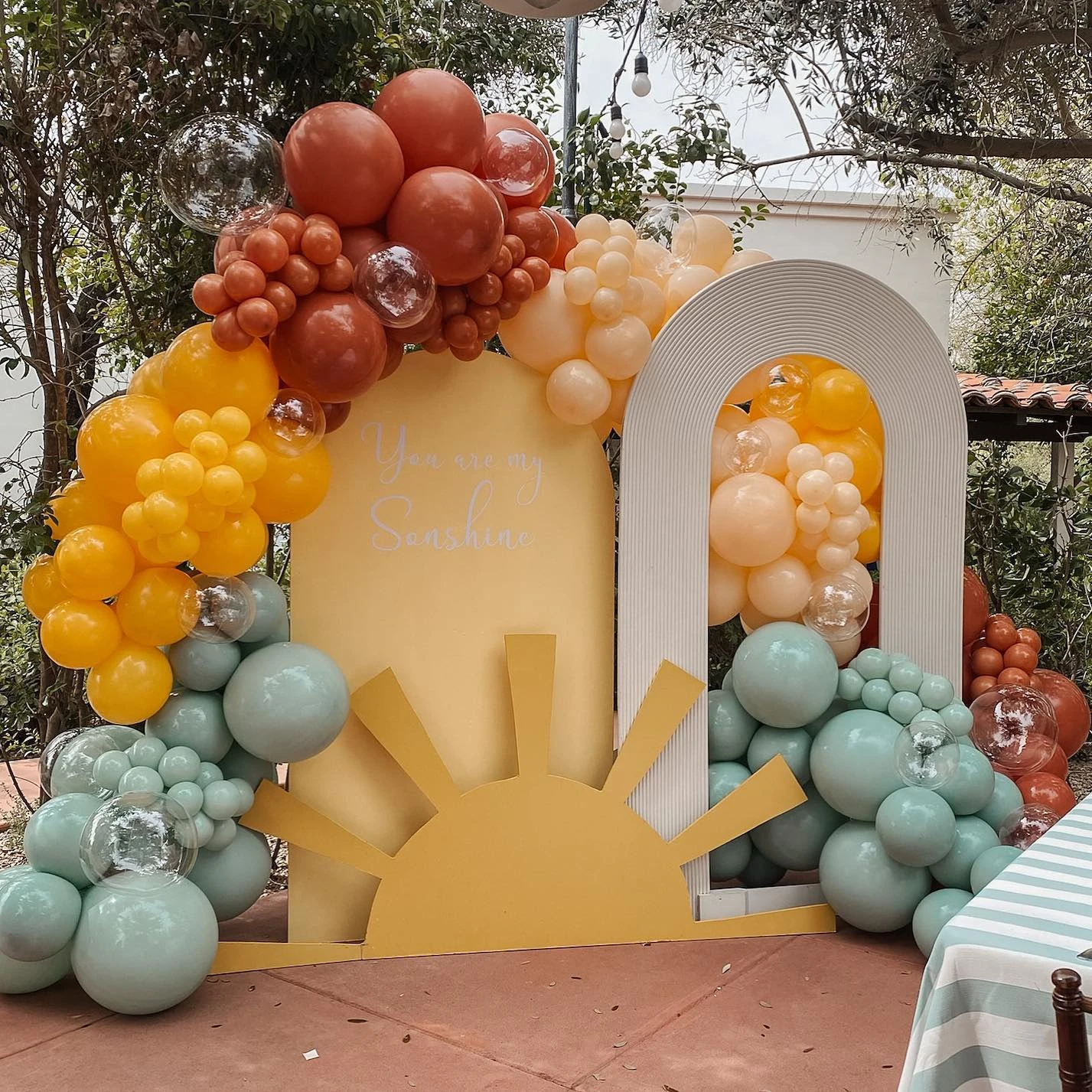 A layered 'You are my sunshine' baby shower backdrop with a large sun cutout and a balloon garland in yellow, orange, and teal.
