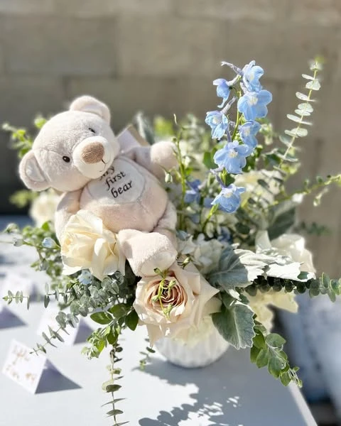 A baby shower centerpiece featuring a cream teddy bear tucked into a white vase with white roses and blue flowers.