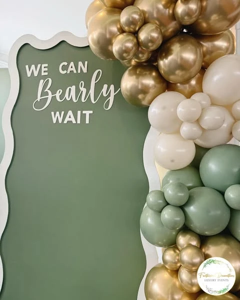 of a 'We Can Bearly Wait' sign in white script lettering on a sage green backdrop, framed by gold and white balloons.
