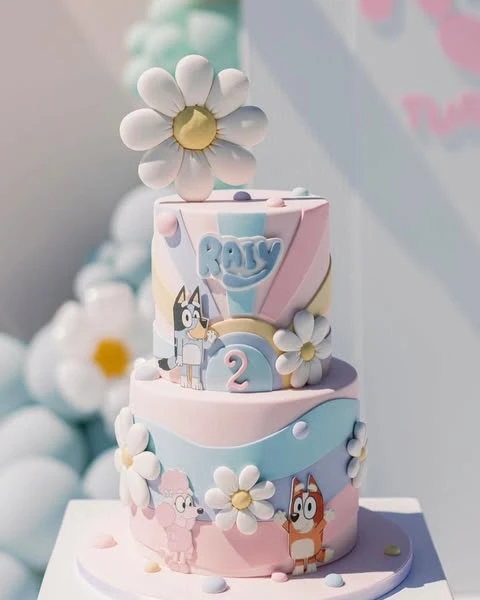 A two-tier Bluey birthday cake decorated in pastel colors with fondant daisies and characters like Bluey, Bingo, and Coco.