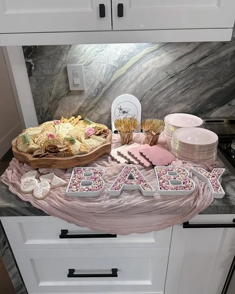 A baby shower food setup featuring letter-shaped dishes spelling 'BABY' filled with candy, next to a platter of crackers and cheese.