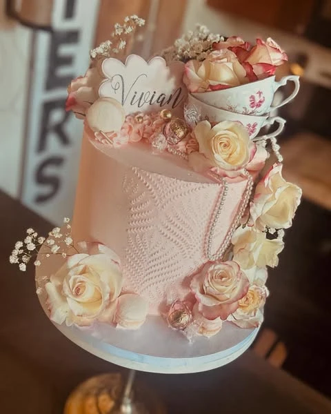 A pink tea party bridal shower cake decorated with a stacked vintage teacup topper and fresh roses.